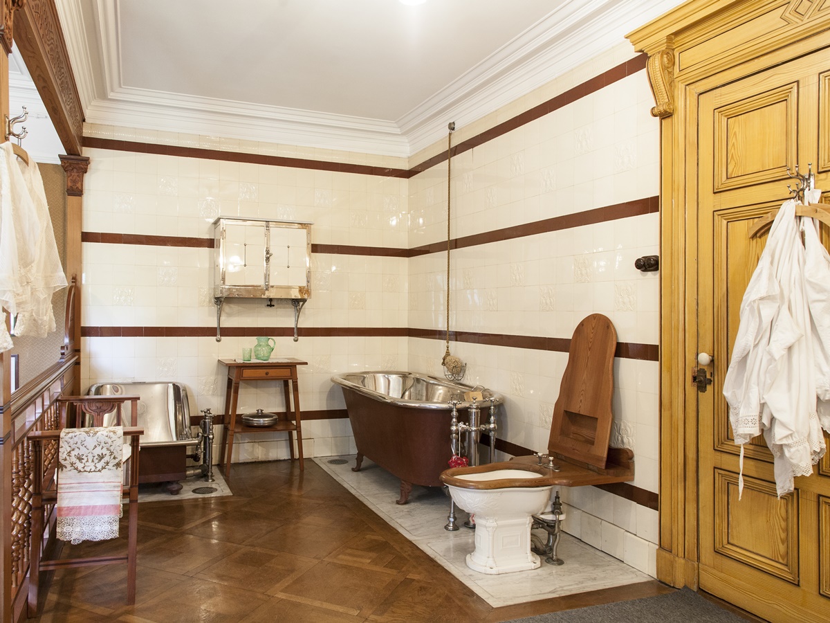 Ein für damalige Verhältnisse luxuriöses Badezimmer im Schloss Hünegg am Thunersee. In der rechten Ecke steht die Badewanne, in der linken Ecke eine Art Sitzbadewanne und mittig davon das Lavabo. Am Fussende der Badewanne die Toilette, die schon fast ganz die heutige Form angenommen hat. Rind und Deckel sind hier noch aus Holz gefertigt. Passend dazu sin an der Türe und Kleiderhaken Textilien der damaligen Zeit aufgehängt.