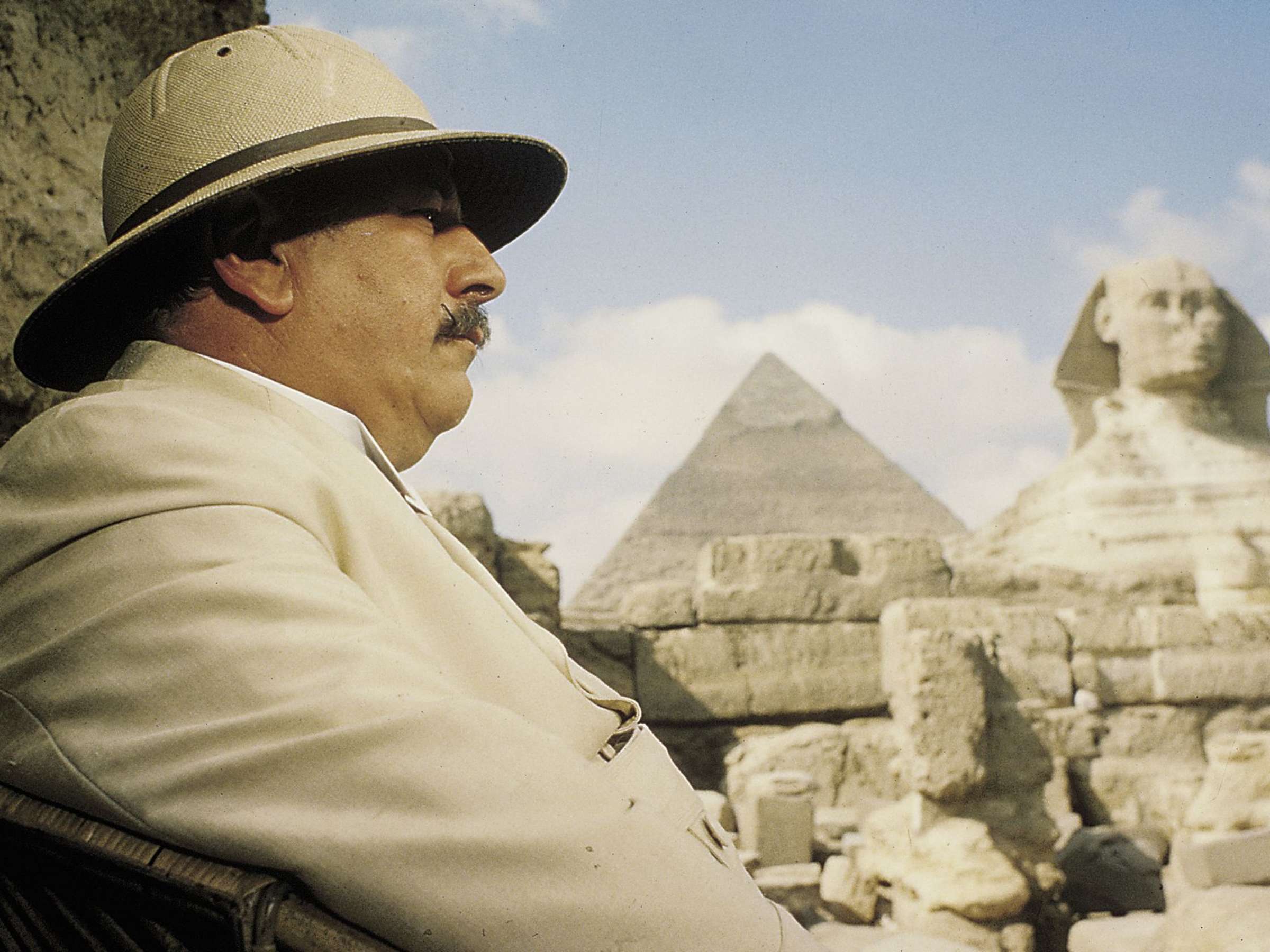 Hercule Poirot sitzt im Schatten einer Pyramide. Rechts im Bild sieht man die Sphinx des alten Ägypten mit der abgebrochenen Nase. Mittig ist eine weitere, weiter entfernte Pyramide zu sehen. Hercule Poirot trägt einen Tropenhut und einen ecrufarbigen Leinen-Anzug.